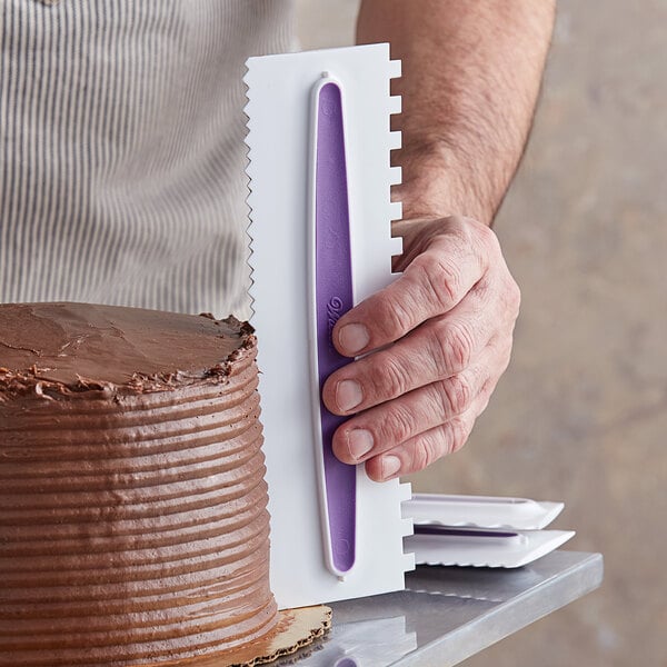 A hand using a Wilton plastic icing comb to cut a chocolate cake.