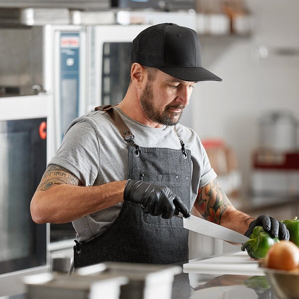 A man in a black Mercer Culinary trucker cap and apron cutting green peppers.