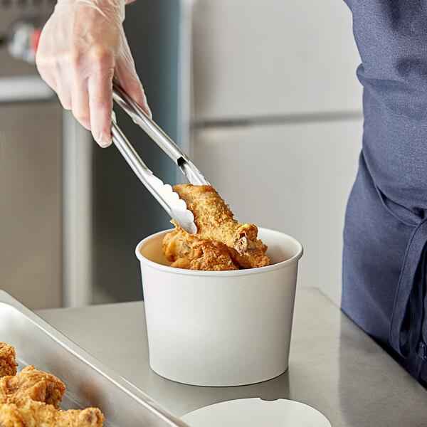 A person using tongs to put fried chicken in a white food bucket.