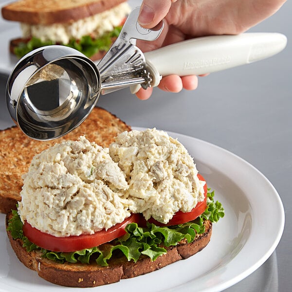A person using a Hamilton Beach ivory metal scoop to put food on a sandwich.
