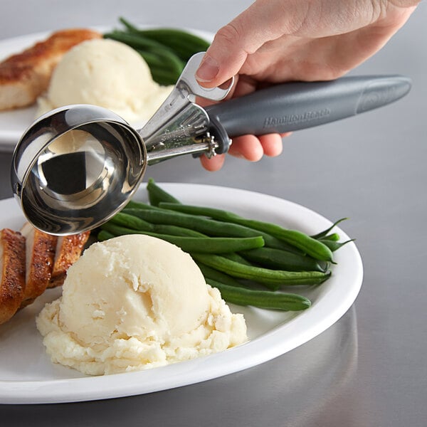 A person holding a Hamilton Beach gray thumb press scooper over a plate of food.