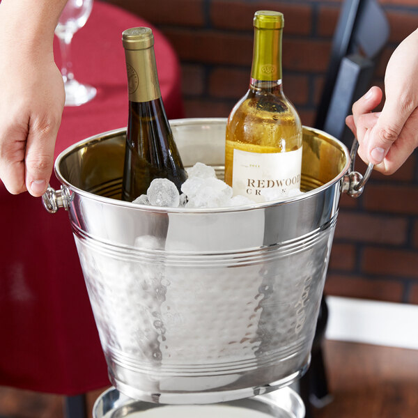 A hand holding a bottle of wine in an American Metalcraft wine bucket filled with ice.