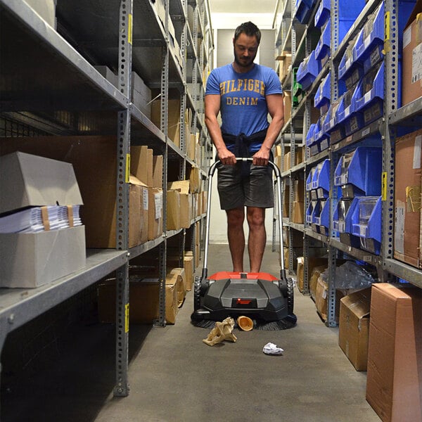 A man in a blue shirt pushing a Bissell Commercial Deluxe Triple Brush Manual Power Sweeper in a warehouse.