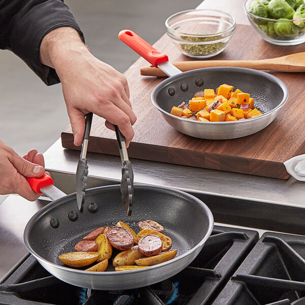 A person cooking food in a Choice aluminum non-stick fry pan with red silicone handles.
