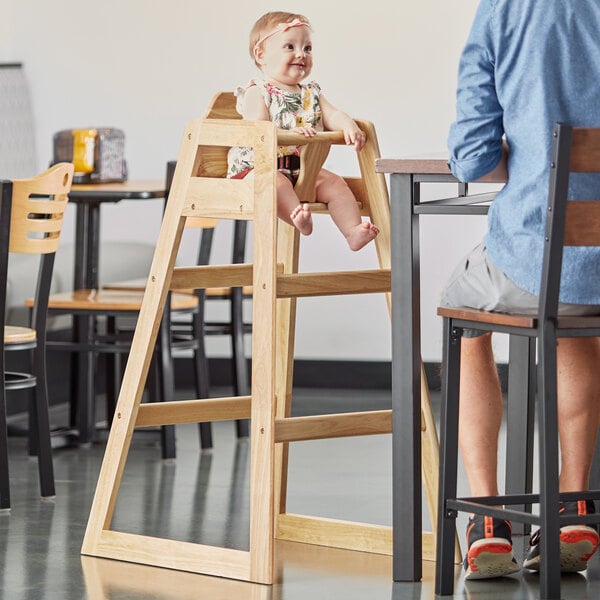 A baby sitting in a Lancaster Table & Seating wooden high chair at a table.