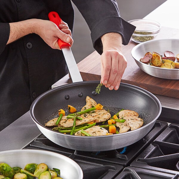 A person cooking food in a Choice aluminum non-stick fry pan with a red silicone handle.