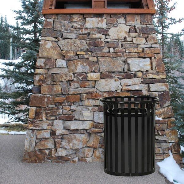 A black Ex-Cell Kaiser Streetscape outdoor trash can next to a stone wall.