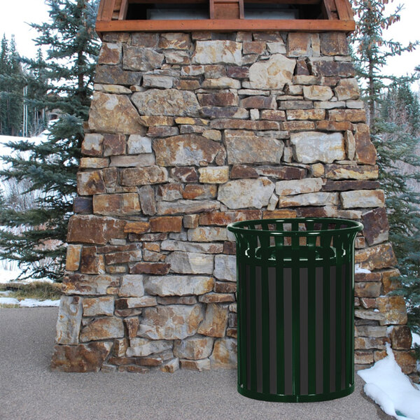 A green Ex-Cell Kaiser Streetscape outdoor trash receptacle next to a stone wall.