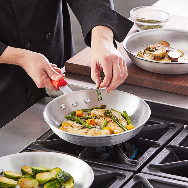 A person cooking chicken and asparagus in a Choice aluminum fry pan with a red silicone handle.