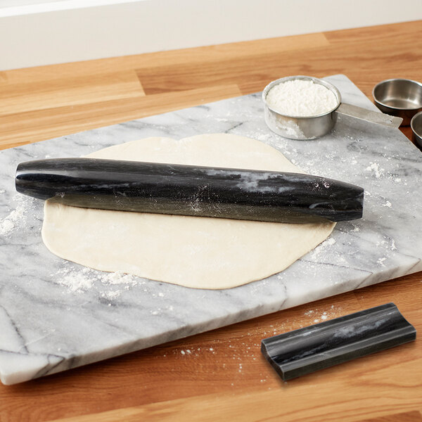 A black marble French rolling pin on a marble surface with flour and measuring cups.