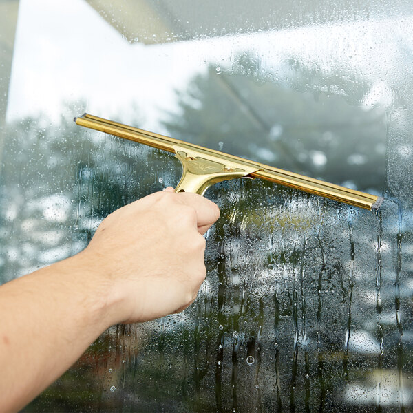 A hand holding a Unger GoldenClip window squeegee with a brass handle to clean a window.