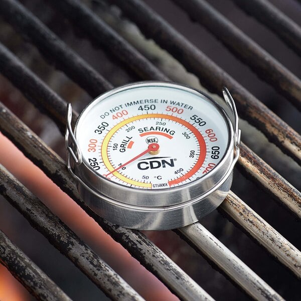 A round metal CDN grill thermometer on a grill.