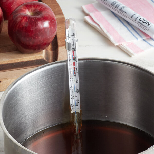A CDN deep fry and candy thermometer in a pot of brown liquid with apples.