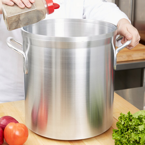 A person pouring seasoning into a silver Vollrath Wear-Ever stock pot.
