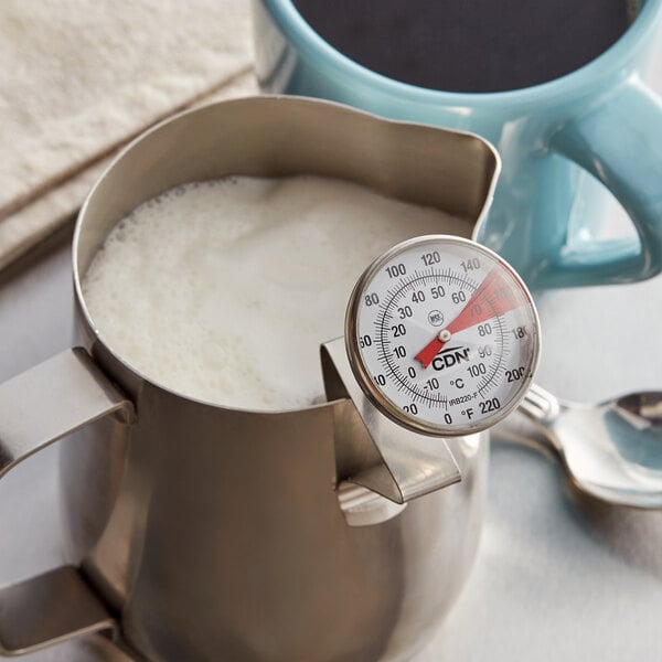 A CDN ProAccurate Insta-Read Frothing Thermometer in a metal container of milk next to a cup of coffee.