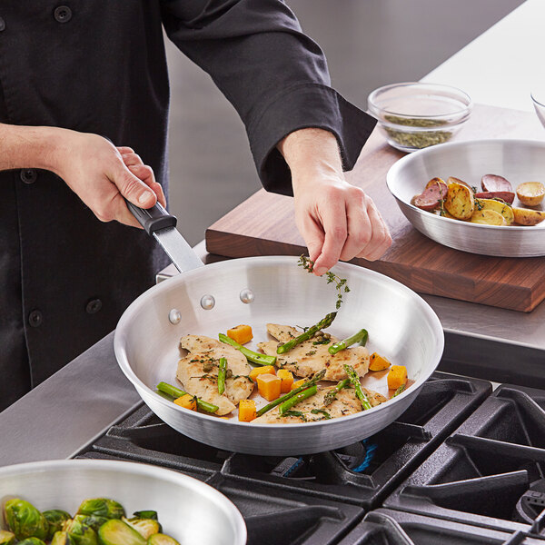 A person cooking chicken and vegetables in a Vollrath aluminum fry pan with a black silicone handle.