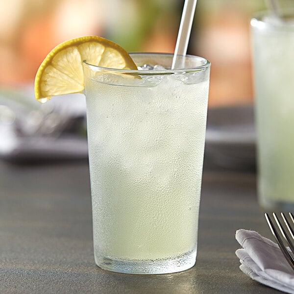 A Duralex glass of lemonade with a straw and ice, with a lemon slice on the table.