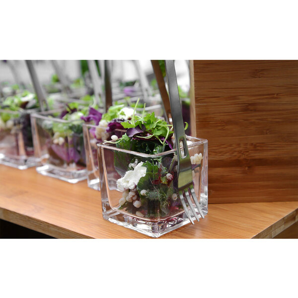 A Front of the House stainless steel taster fork in a salad in a square glass container.
