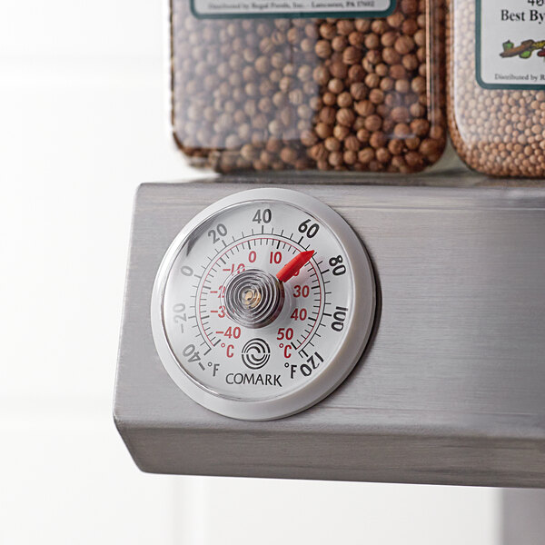 A Comark stick-on thermometer on a metal shelf with food in containers.
