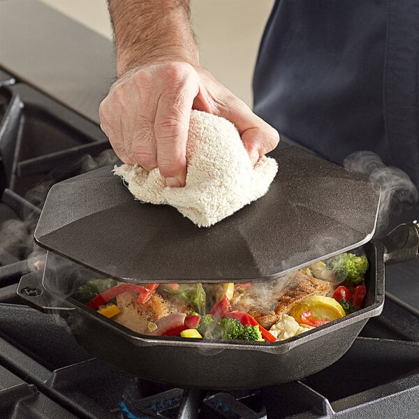 A person's hand holding a white towel while using a FINEX octagonal cast iron cover on a skillet over a stove.