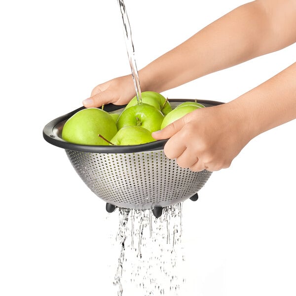 A person using an OXO stainless steel colander to wash green apples.