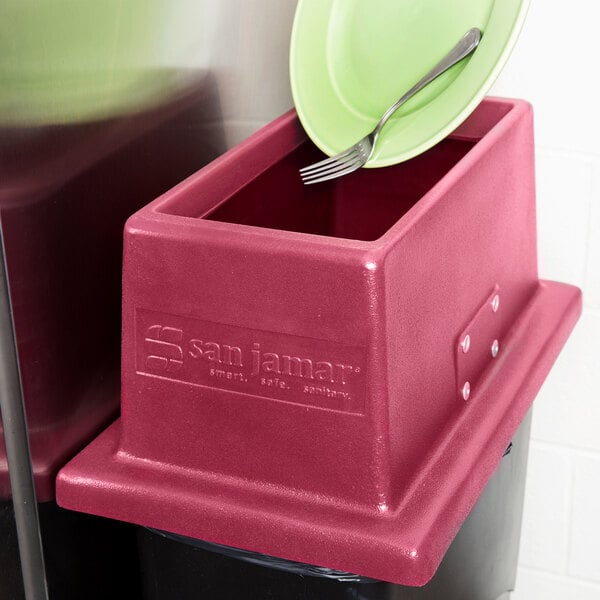 A red San Jamar KatchAll flatware retriever lid on a Slim Jim trash can with a fork on a green plate.