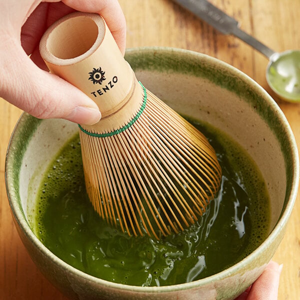 A hand using a Tenzo bamboo whisk to stir green matcha in a bowl.