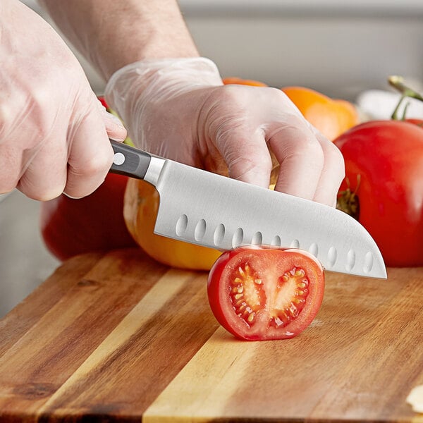 A person in gloves using a Wusthof Classic Santoku knife to cut a tomato on a wooden surface.