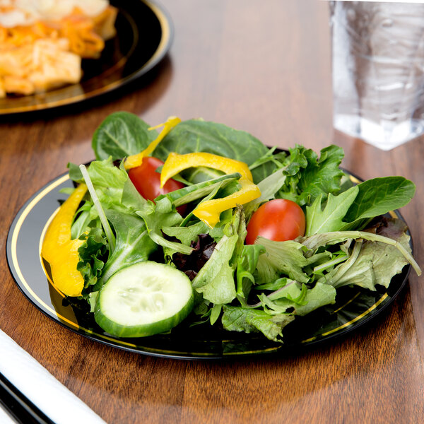 A Fineline Silver Splendor black plastic plate with gold bands holding a salad on a table.