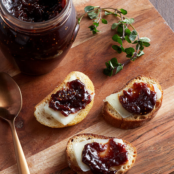A wooden cutting board with bread and Dalmatia Fig Cocoa spread on it, next to a spoon and a jar of jam.
