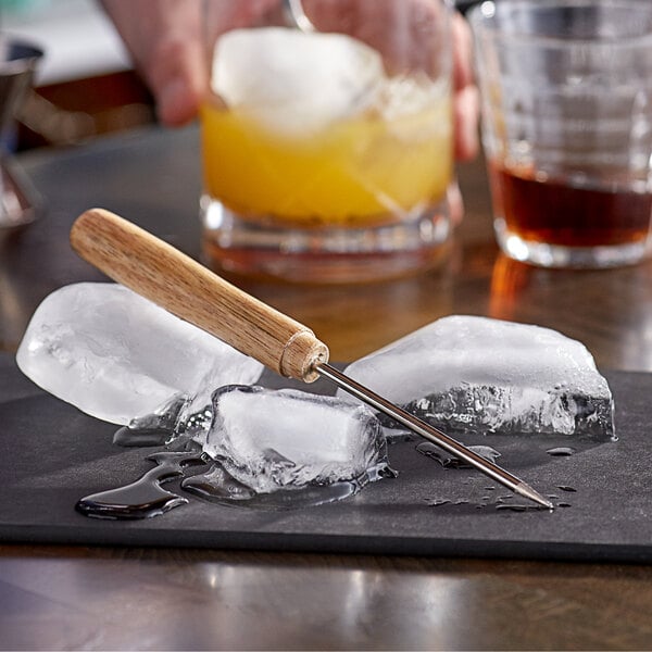 An American Metalcraft steel ice pick with a wooden handle on a black surface with ice cubes.