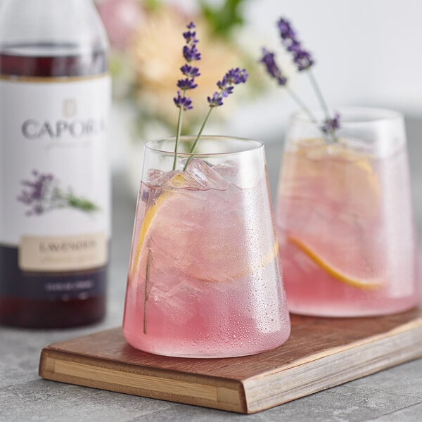 Two glasses of pink lavender lemonade with lemon slices and lavender flowers on a table with a bottle of Capora Lavender Flavoring Syrup.