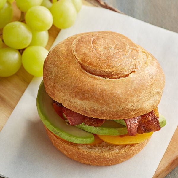 An unsliced Upper Crust croissant bun on a cutting board with a sandwich and fruit.