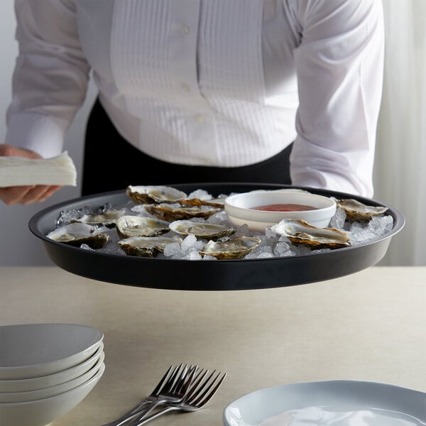 A person in a white shirt holding a charcoal plastic serving tray of oysters on ice.