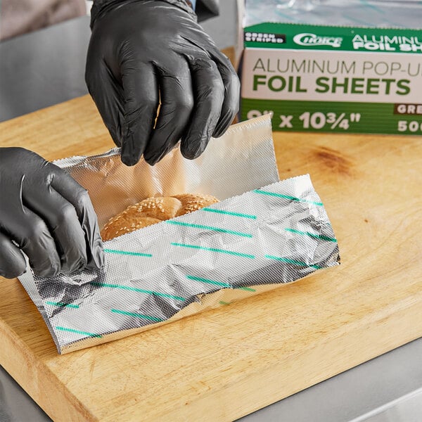 A person in black gloves using Choice green striped interfolded foil to wrap bread.