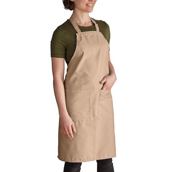 A woman wearing an Intedge beige poly-cotton bib apron with pockets.