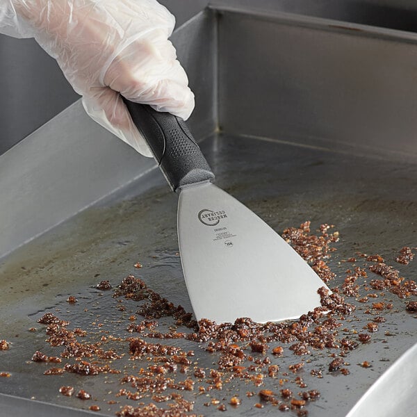 A hand in a glove holding a Mercer Culinary High Carbon Stainless Steel Grill Scraper with a black handle.