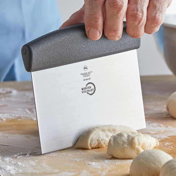 A person using a Mercer Culinary stainless steel bench scraper to cut white dough.