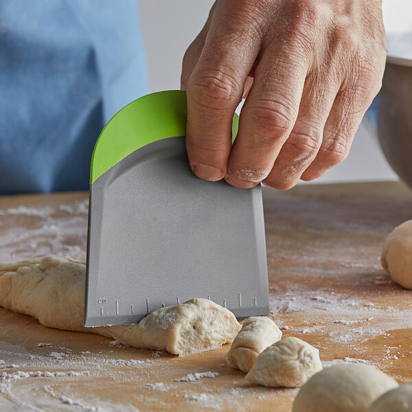 A person using a Fox Run gray plastic dough cutter with a green silicone handle to cut dough.