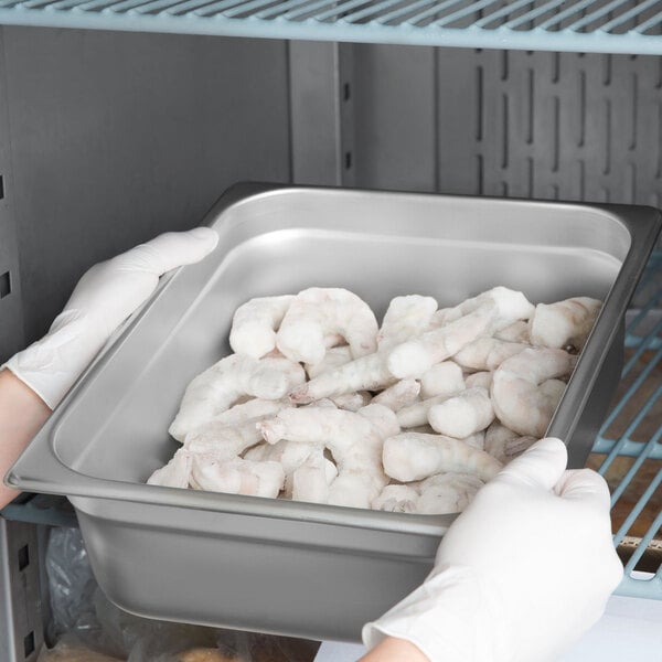 A person in gloves holding a grey stainless steel steam table pan filled with shrimp.