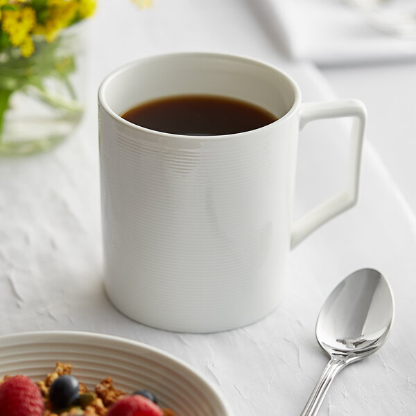A white Acopa Liana porcelain mug filled with brown liquid on a table with a bowl of cereal and berries.