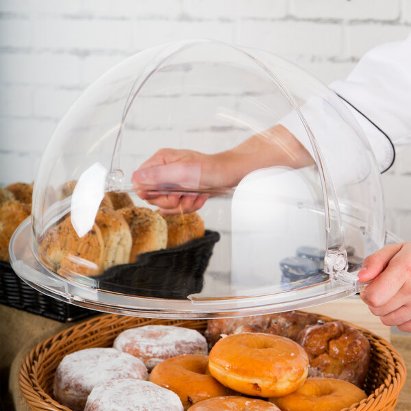 A person holding a GET Designer Polyweave clear round cover over a tray of donuts in a bakery display.