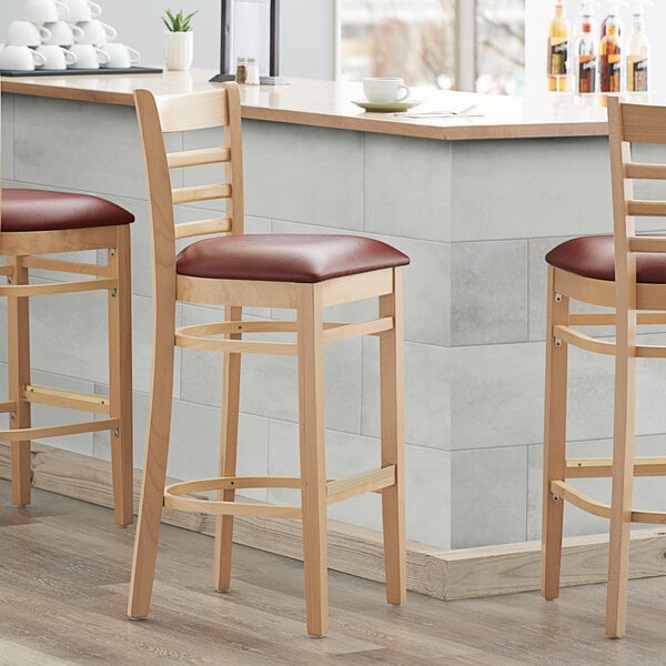 Three Lancaster Table & Seating wood ladder back bar stools with burgundy vinyl seats at a counter.