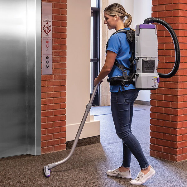 A woman wearing a blue shirt using a ProTeam cordless backpack vacuum to clean a room.