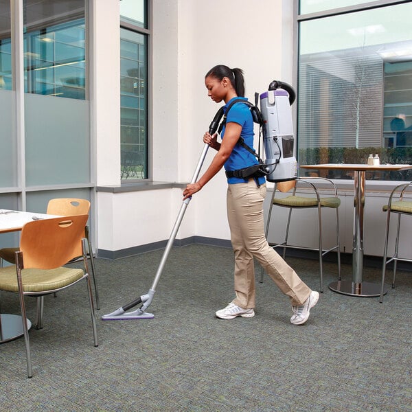 A woman using a ProTeam GoFree Flex Pro II cordless backpack vacuum to clean the floor of a corporate office cafeteria.
