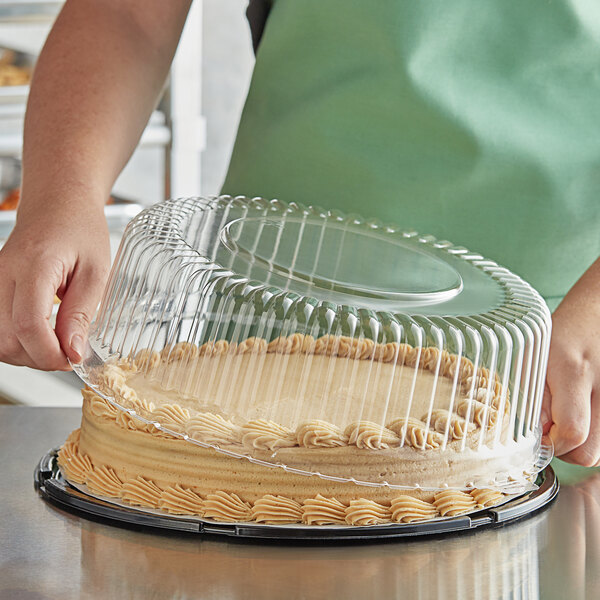 A person placing a cake with frosting into a Baker's Mark cake display container with a clear plastic lid.