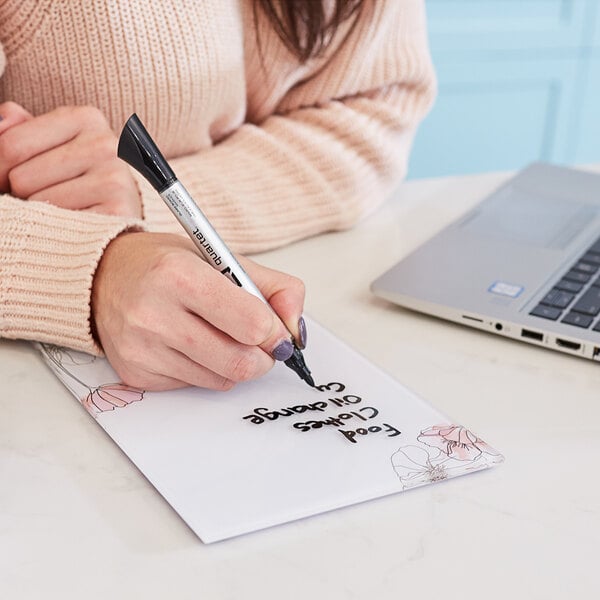A person using a Quartet white glass dry erase notepad to write on a piece of paper on a table.