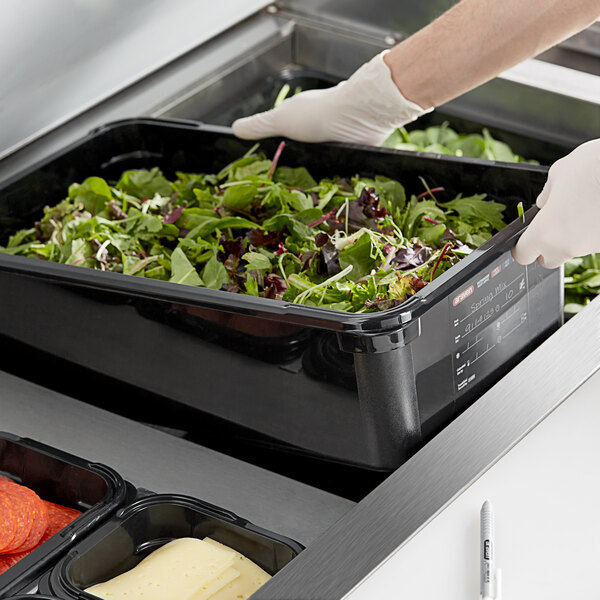 A hand in a glove holding an Araven black plastic food pan filled with salad.