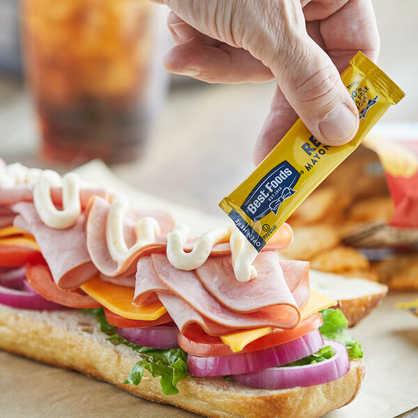 A hand holding a Best Foods mayonnaise packet over a sandwich.