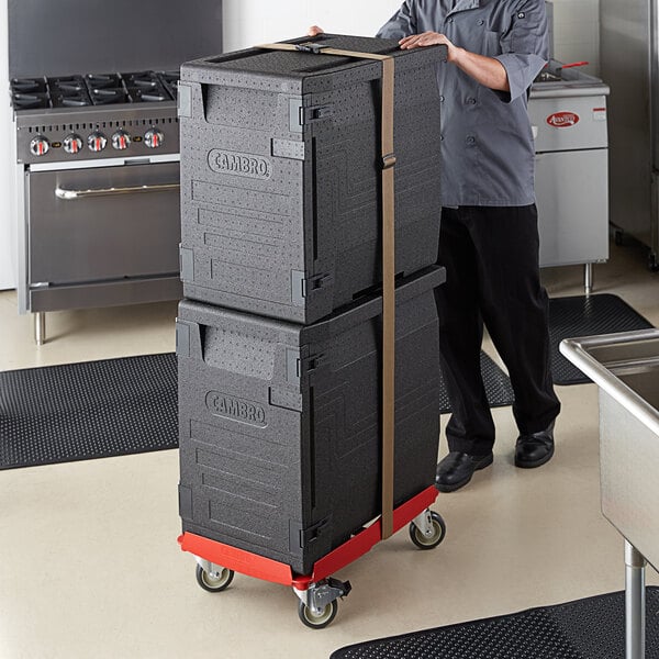 A man pushing a stack of black Cambro Cam GoBox pan carriers on a red and white Camdolly.